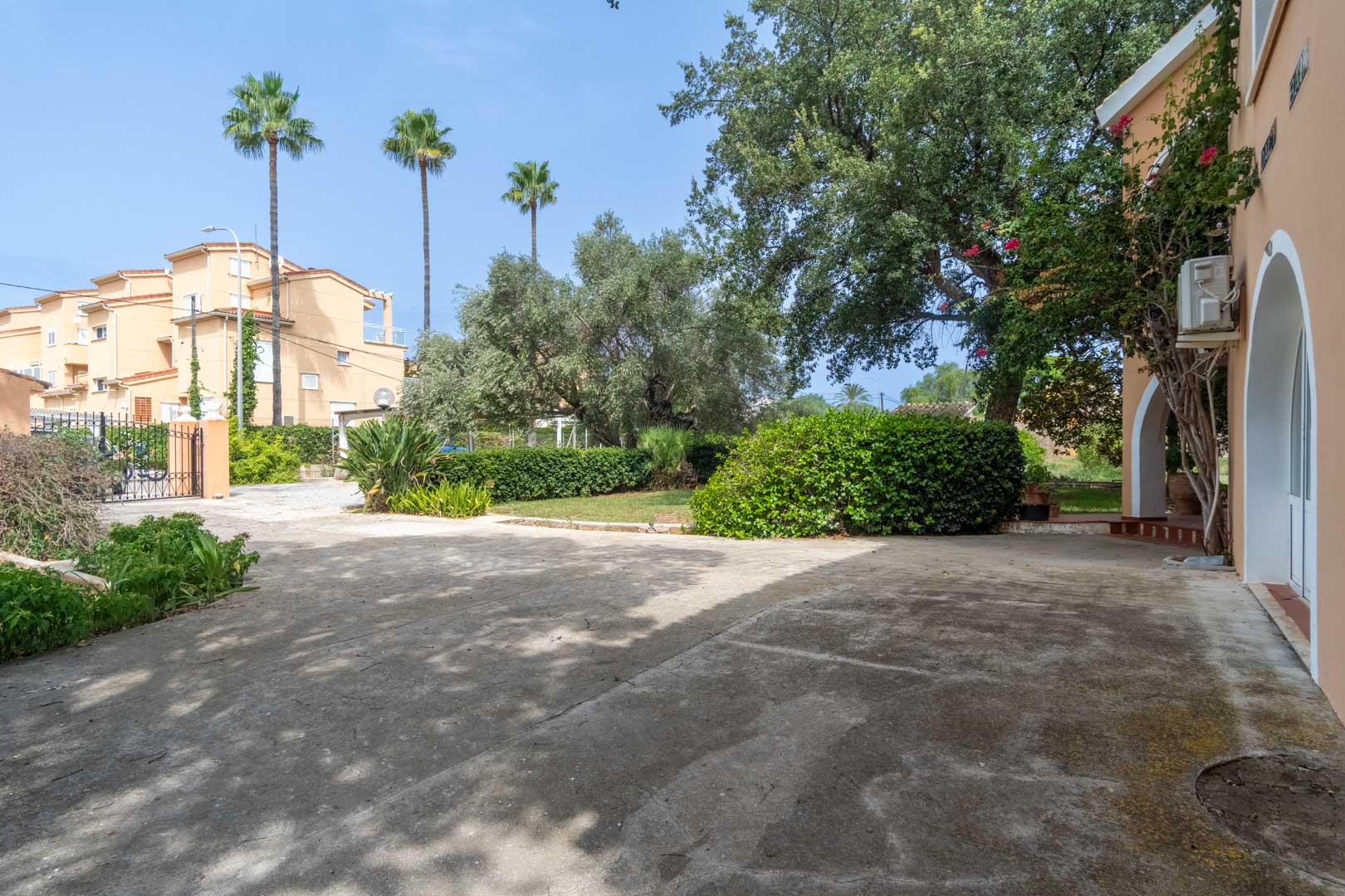Amplia zona exterior pavimentada con acceso mediante puerta de forja, rodeada de vegetación mediterránea, palmeras y fachada con arcos tradicionales.