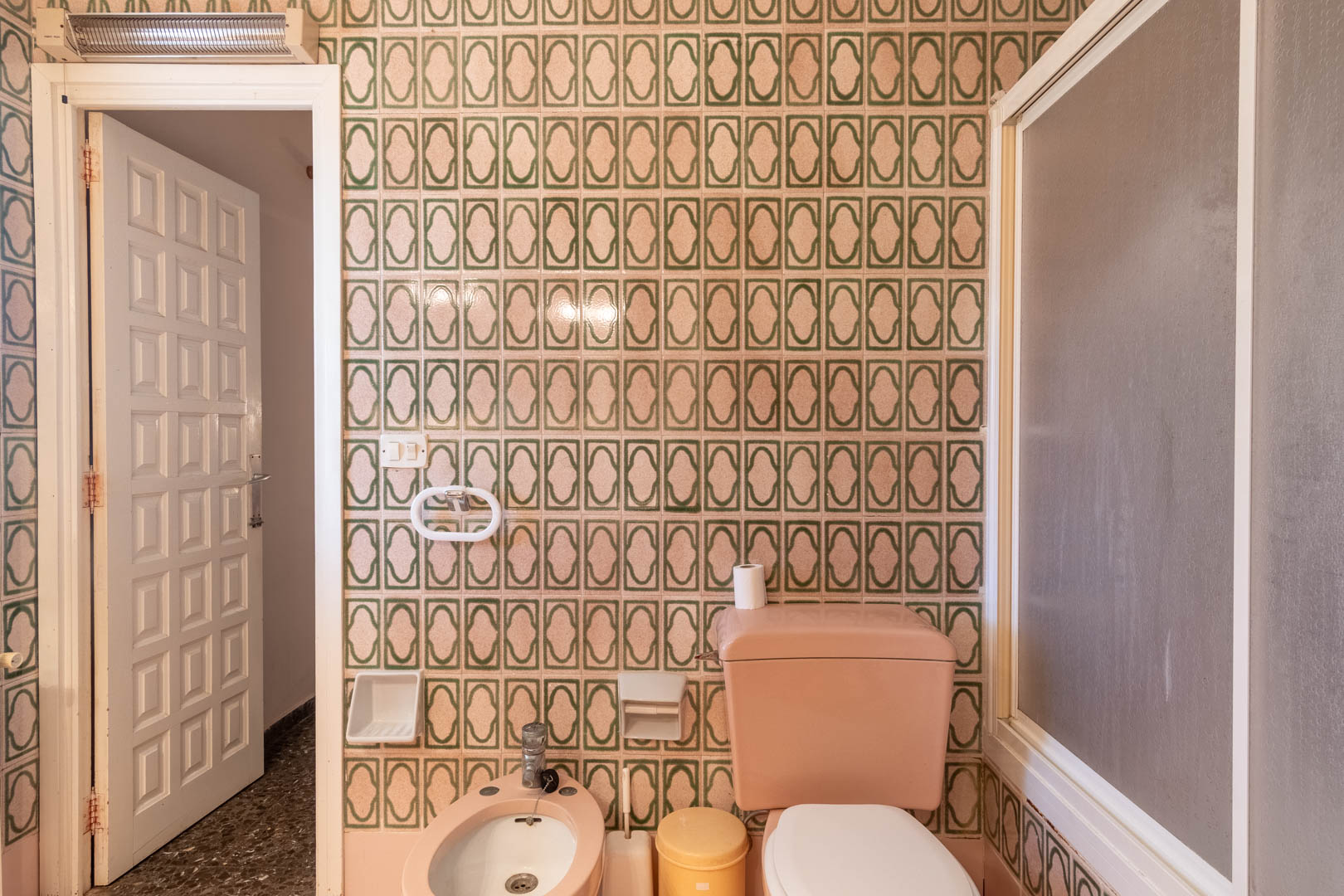 Baño con azulejos de patrón geométrico verde, sanitarios de porcelana rosa que incluyen bidet e inodoro, y una puerta blanca de cuarterones.