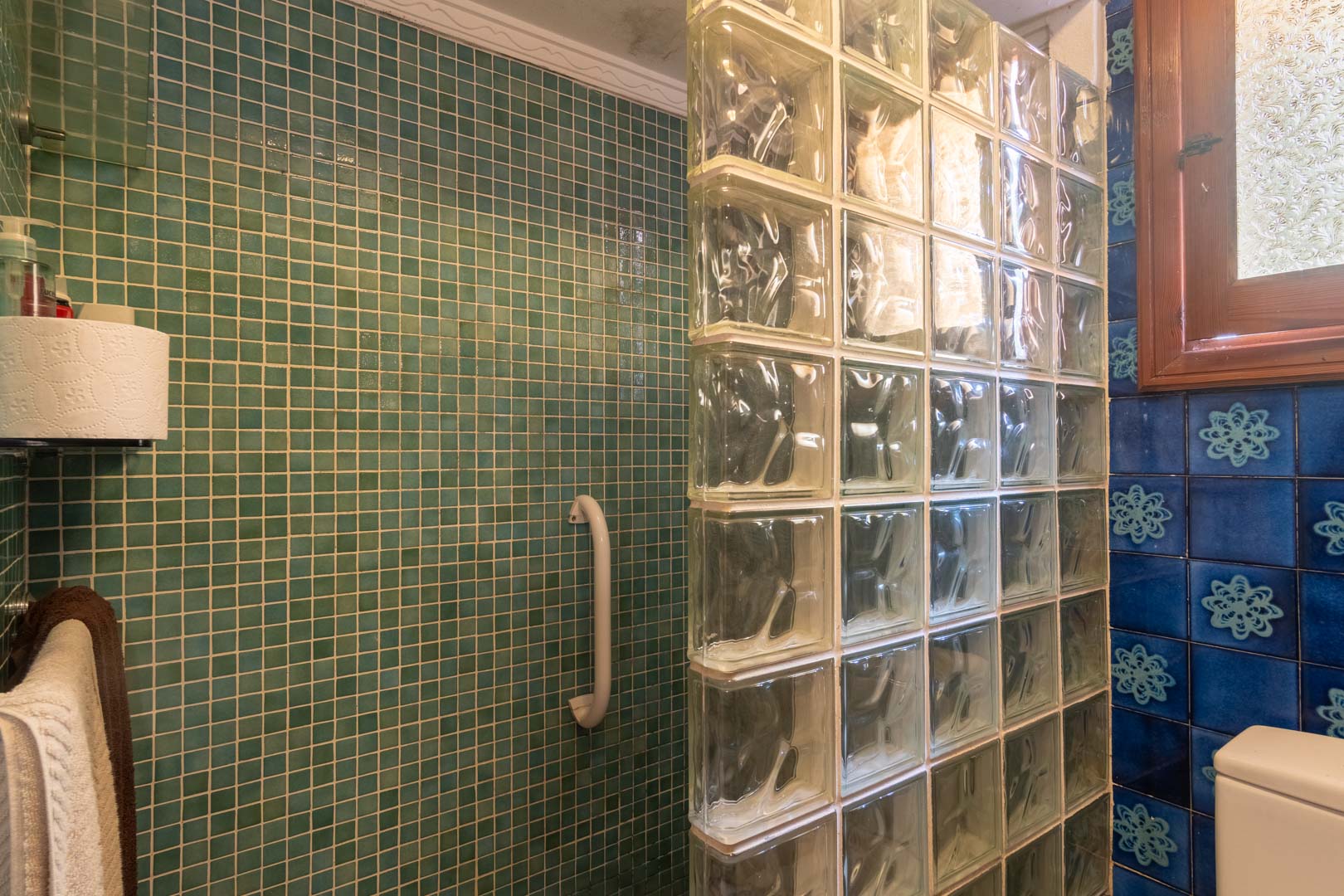 Baño con zona de ducha en mosaico verde y tabique de bloques de vidrio. Presenta azulejos azules con motivos florales y una ventana de madera que aporta luz natural.