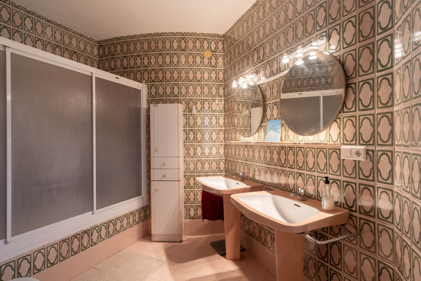 Baño de estilo retro con lavabos de doble pedestal, espejos circulares iluminados y azulejos con motivos verdes. Incluye ducha con mampara translúcida y armario de almacenaje.
