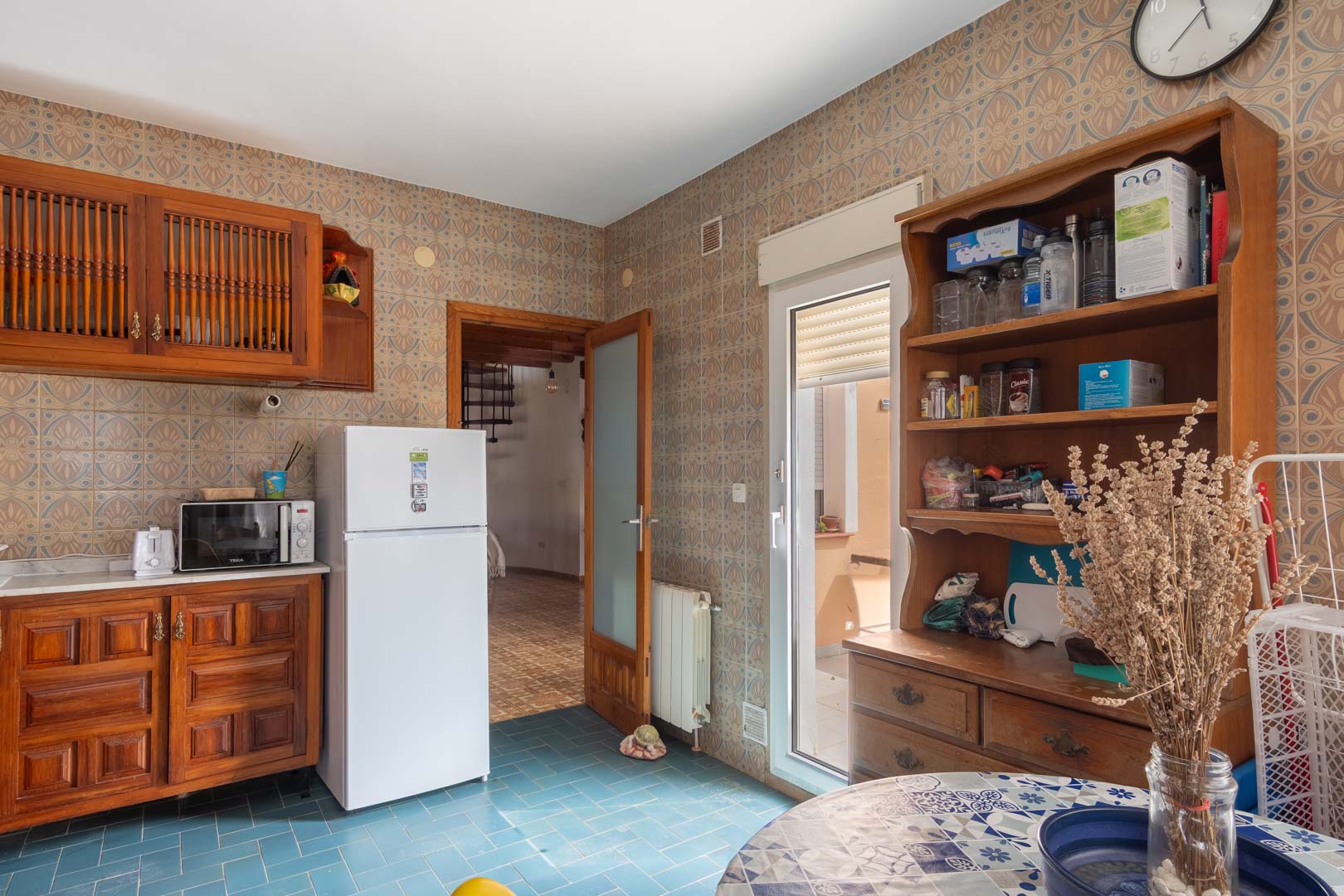 Cocina de estilo rústico con armarios de madera, suelos cerámicos azules y paredes decoradas. Incluye acceso directo al exterior y vistas al salón contiguo con escalera.
