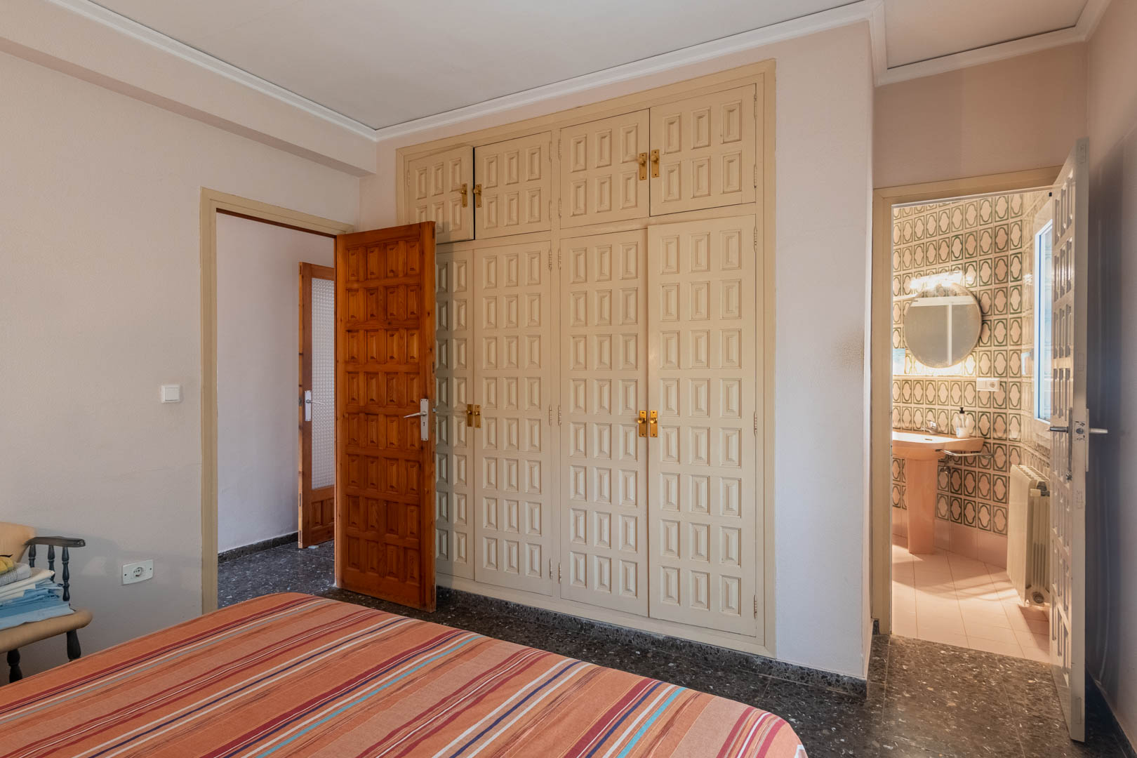 Dormitorio con amplio armario empotrado de paneles decorativos y acceso directo a un baño en suite con azulejos de diseño geométrico y luz natural.