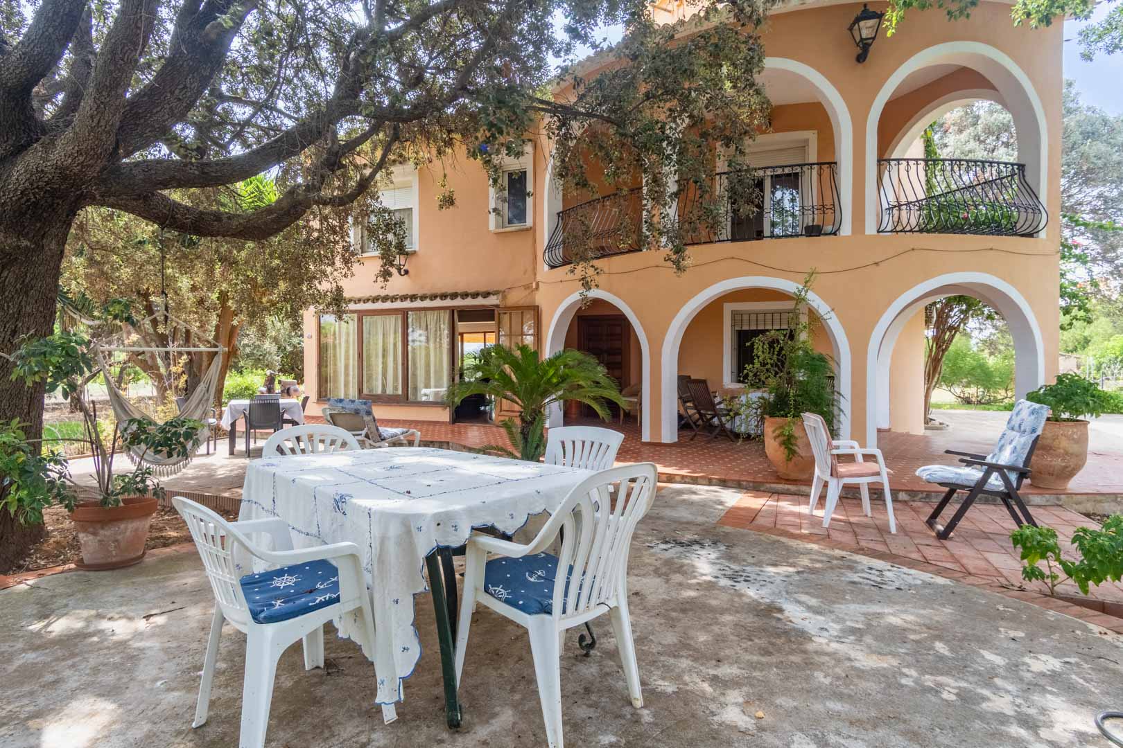 Fachada mediterránea con porches de arcos blancos, amplias terrazas sombreadas por árboles maduros y zonas de comedor exterior sobre suelo de barro.