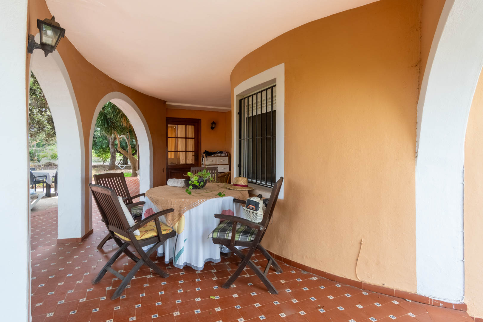 Porche cubierto con arcos blancos y suelos de gres rústico. Incluye zona de comedor exterior protegida del sol con vistas directas al jardín mediterráneo.
