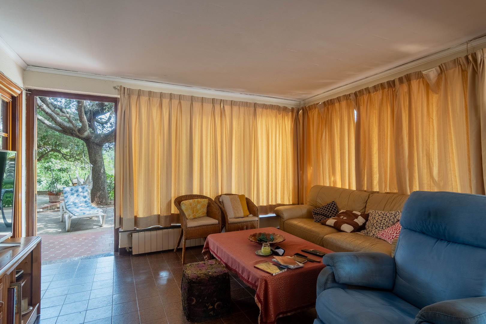 Salón con suelo cerámico, mobiliario variado y acceso directo a una terraza sombreada por un gran árbol. Los ventanales cuentan con cortinas de tonos cálidos y radiadores.