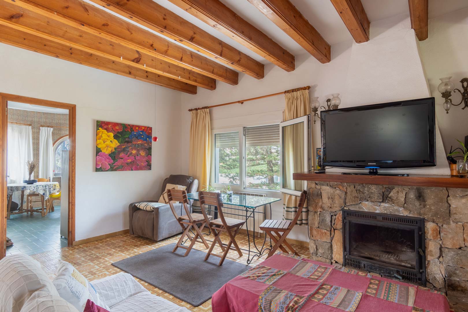 Salón rústico con vigas de madera a la vista, chimenea de piedra natural y zona de comedor con mesa de cristal bajo luz natural.