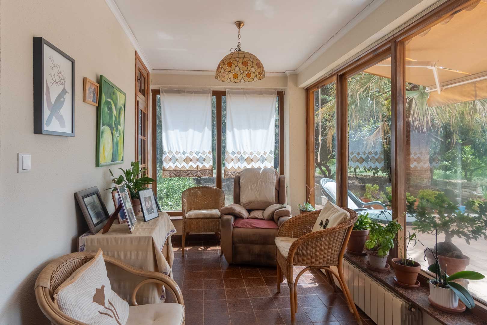 Terraza acristalada con carpintería de madera y suelos cerámicos. Un espacio luminoso con vistas al jardín, mobiliario de mimbre y una atmósfera acogedora.
