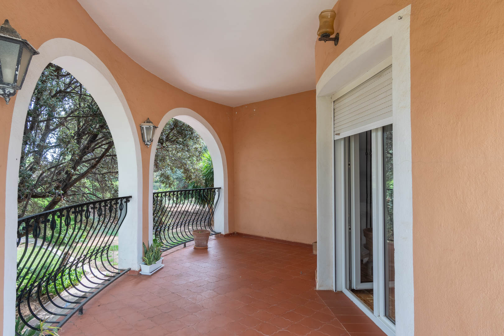 Terraza cubierta con arcos de estilo mediterráneo, suelo de baldosas cerámicas y barandilla de hierro forjado con vistas al jardín arbolado.