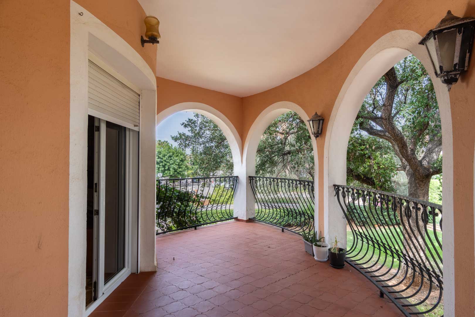Terraza cubierta con arcos de medio punto, barandilla de hierro forjado curvada y suelo de baldosas cerámicas con vistas al jardín mediterráneo.