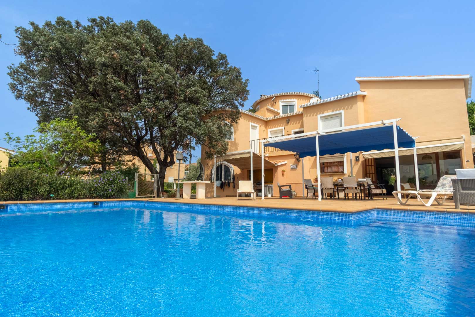 Vista exterior de la piscina privada rodeada de una amplia terraza de gres, con el chalet mediterráneo y su zona de comedor bajo toldo azul al fondo.