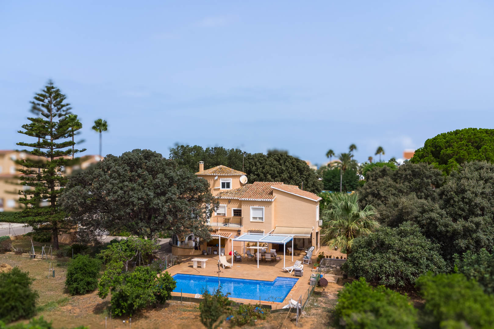 Vista exterior del chalet con piscina rectangular, terraza con pérgola blanca y zona de comedor al aire libre rodeada de vegetación mediterránea y árboles maduros.
