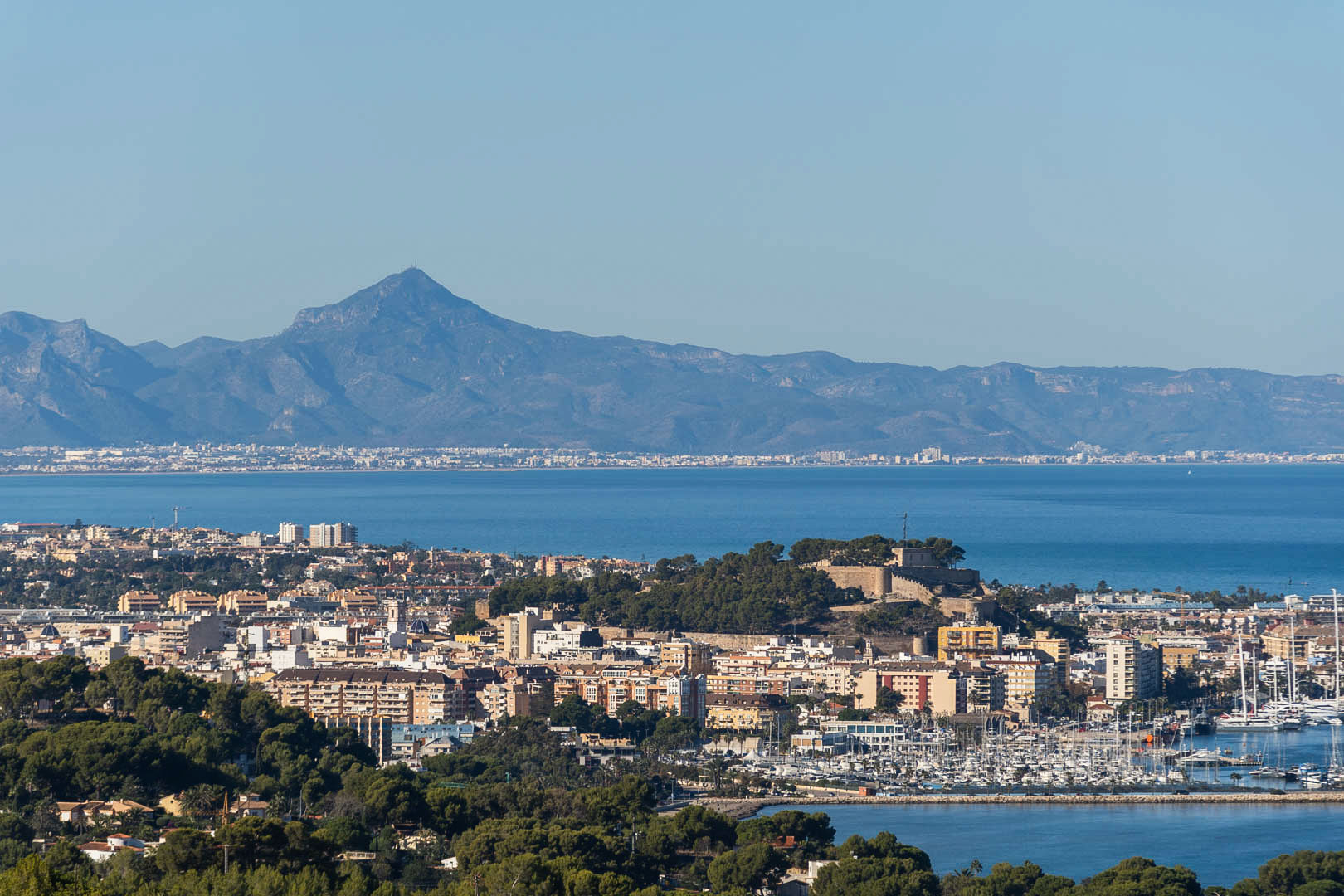 Vista panorámica de Dénia que muestra el castillo histórico, el puerto deportivo con sus embarcaciones y la silueta de las montañas frente al mar Mediterráneo.