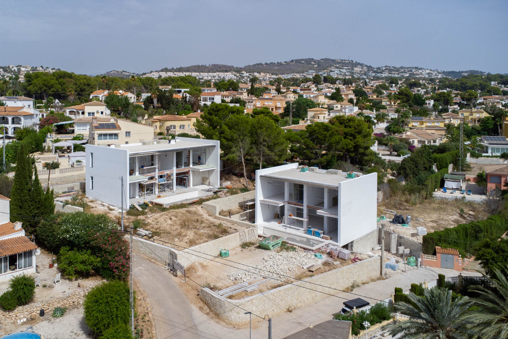 Vista aérea de la fase de construcción de dos villas modernas en Calpe, mostrando las estructuras blancas, muros de piedra natural y la nivelación de las parcelas.