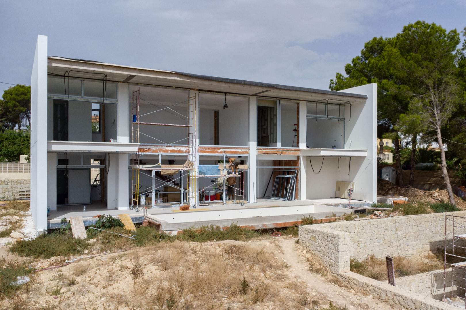Vista exterior de la villa en construcción con estructura de dos niveles, amplios marcos para ventanales y la zona de la piscina en proceso.