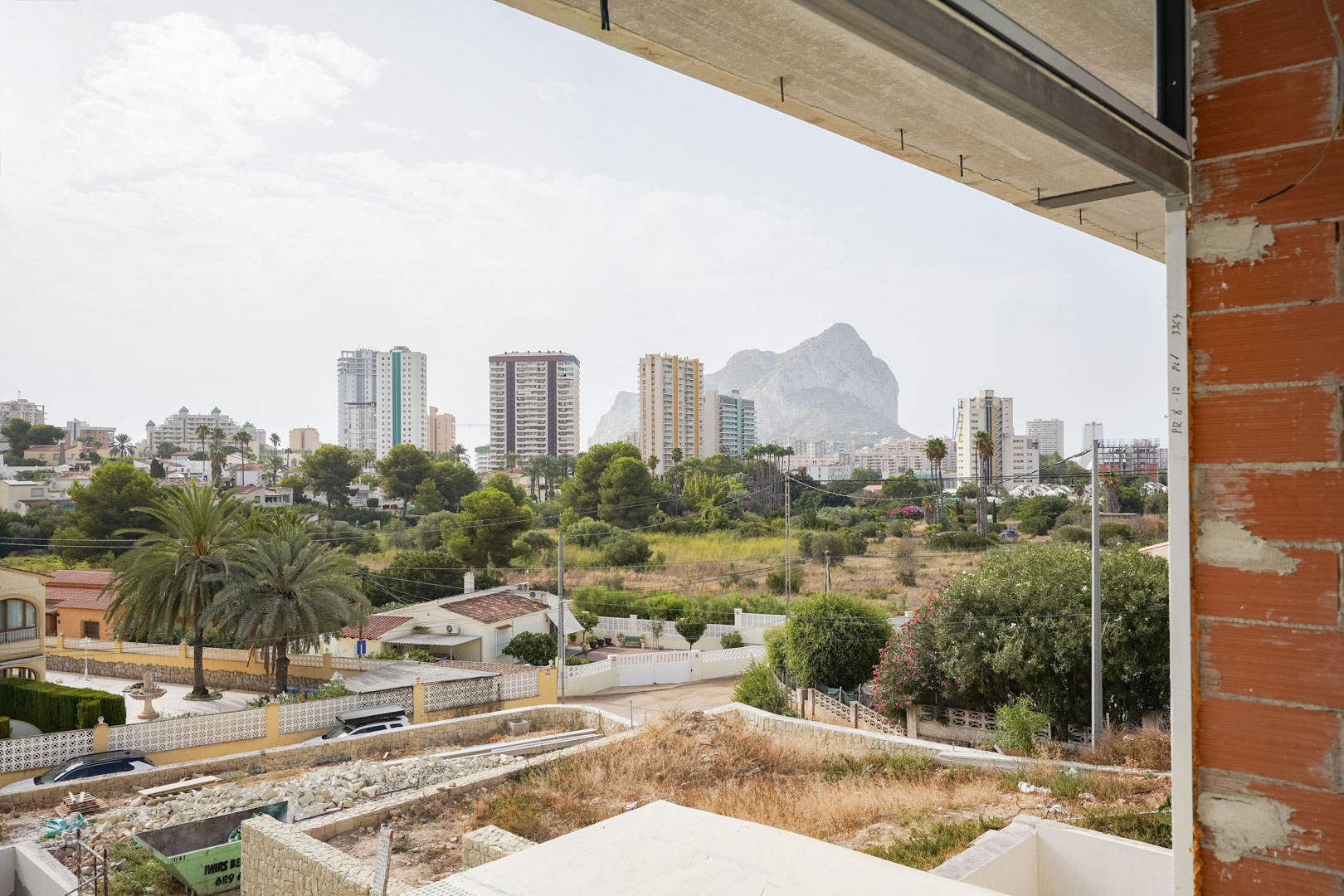 Vista panorámica desde la planta superior en construcción hacia el Peñón de Ifach y el skyline de Calpe, enmarcada por muros de ladrillo y forjados de hormigón.