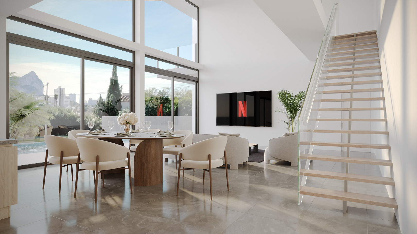 Salón-comedor de doble altura con grandes ventanales, vistas al Peñón de Ifach, suelos de piedra pulida y una escalera flotante de madera con barandilla de cristal.