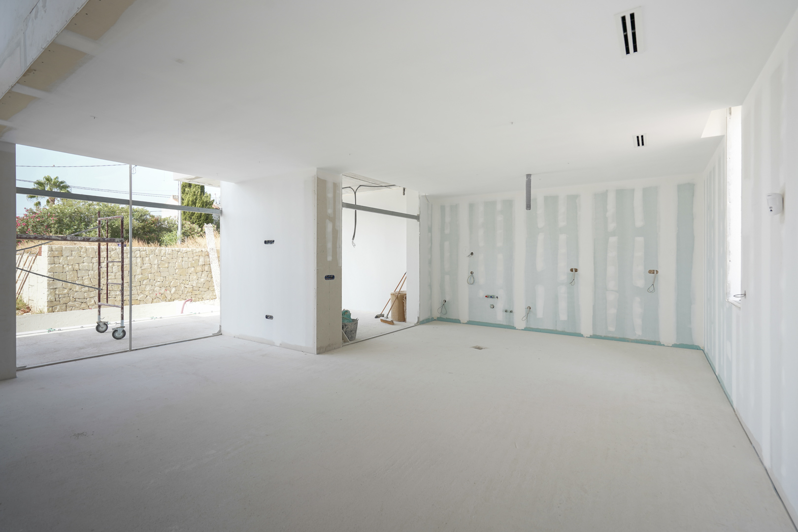 Vista interior del salón en construcción con conductos de aire acondicionado integrados y amplios ventanales hacia la zona de terraza y jardín.
