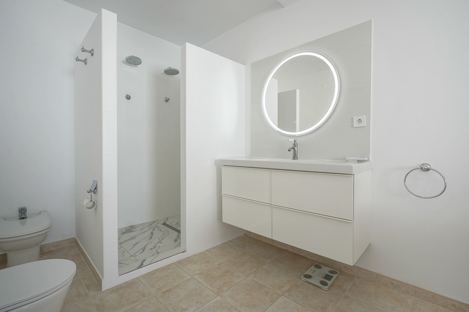 Baño moderno con mueble suspendido blanco, espejo circular retroiluminado y una amplia zona de ducha doble con platos de efecto mármol y grifería empotrada.