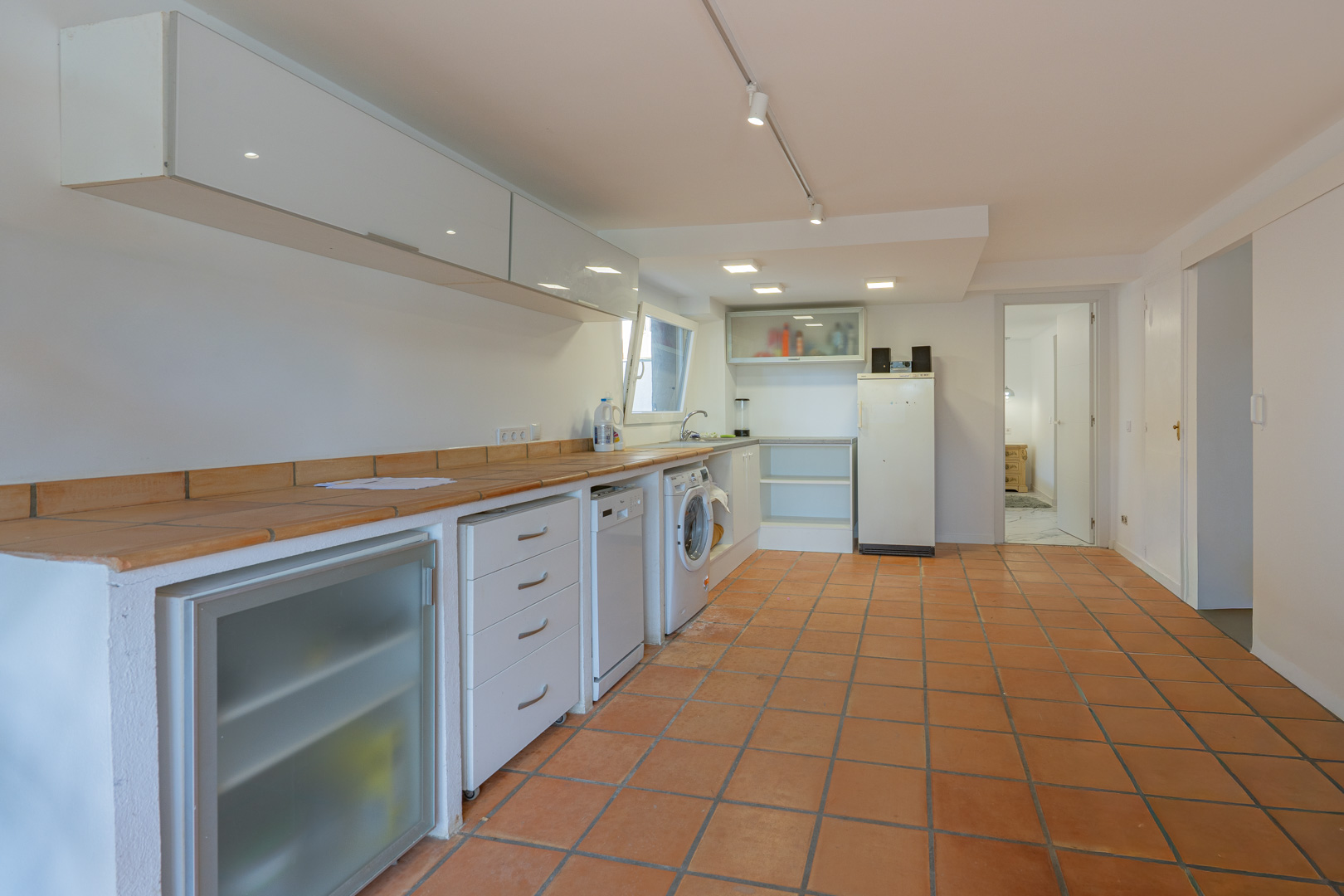 Cocina adicional y zona de lavandería con suelos de terracota, encimera de azulejo y electrodomésticos integrados bajo armarios blancos minimalistas.