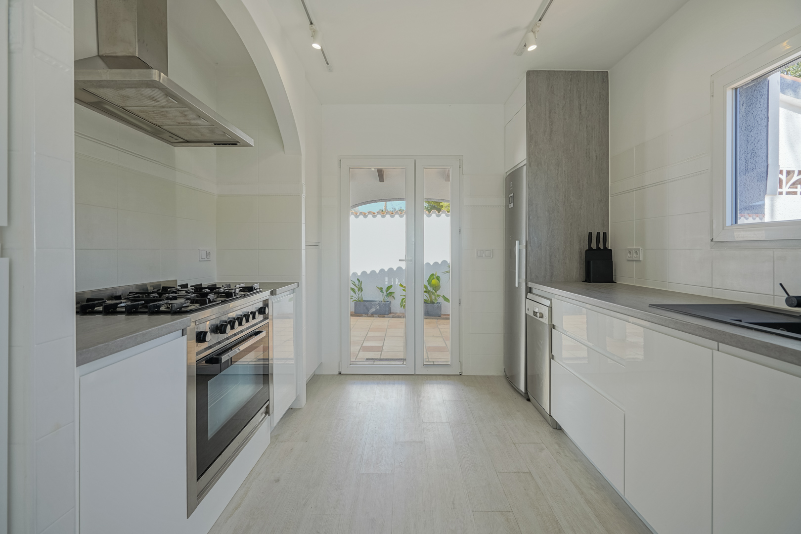 Cocina moderna con mobiliario blanco brillante, encimeras grises y electrodomésticos de acero inoxidable. Incluye cocina de gas y acceso directo a la terraza exterior.