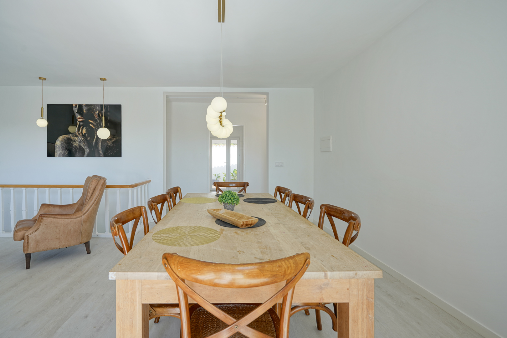 Comedor con mesa de madera clara y sillas clásicas, iluminado por lámparas colgantes esféricas. Al fondo se aprecia un sillón de cuero y una pintura artística sobre paredes blancas.