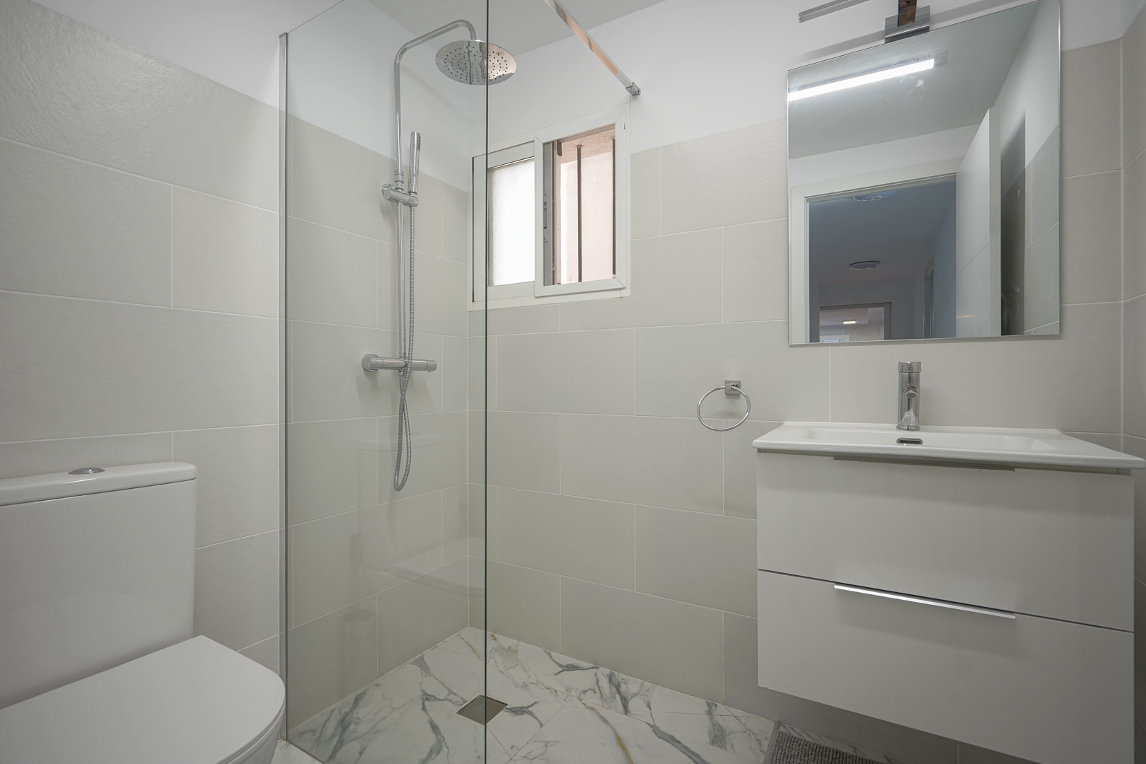 Cuarto de baño moderno con ducha a ras de suelo, mampara de cristal, lavabo con mueble blanco y suelos cerámicos con efecto mármol.