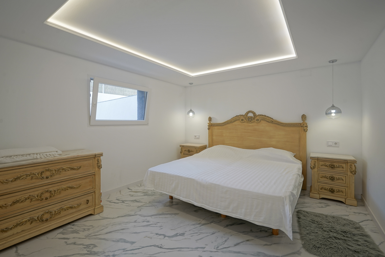 Dormitorio con mobiliario clásico tallado, suelos de gres porcelánico efecto mármol y techo con iluminación LED perimetral integrada.