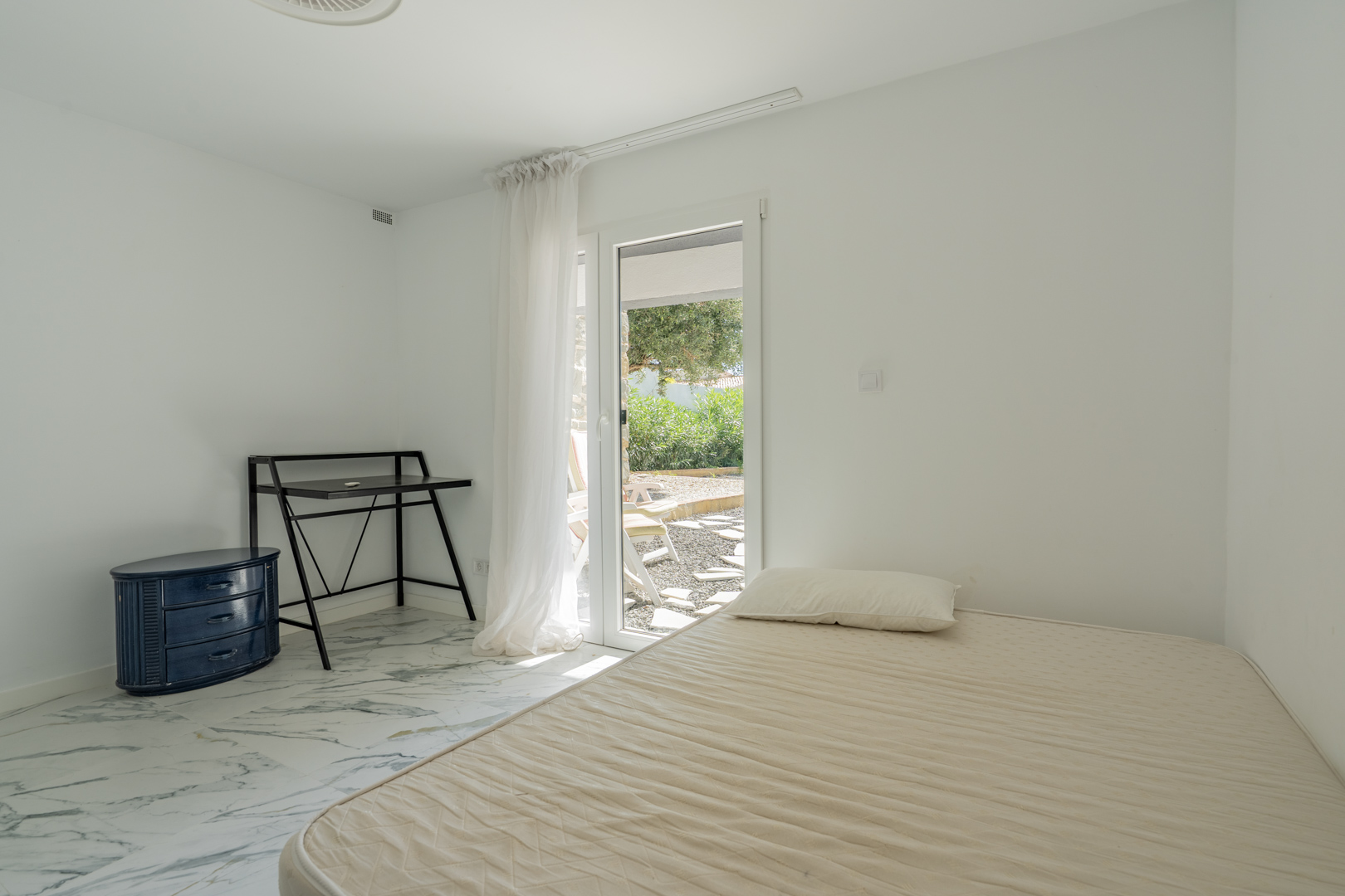 Dormitorio con suelos de mármol y acceso directo al jardín a través de una puerta acristalada. Incluye un escritorio funcional y cómoda azul.