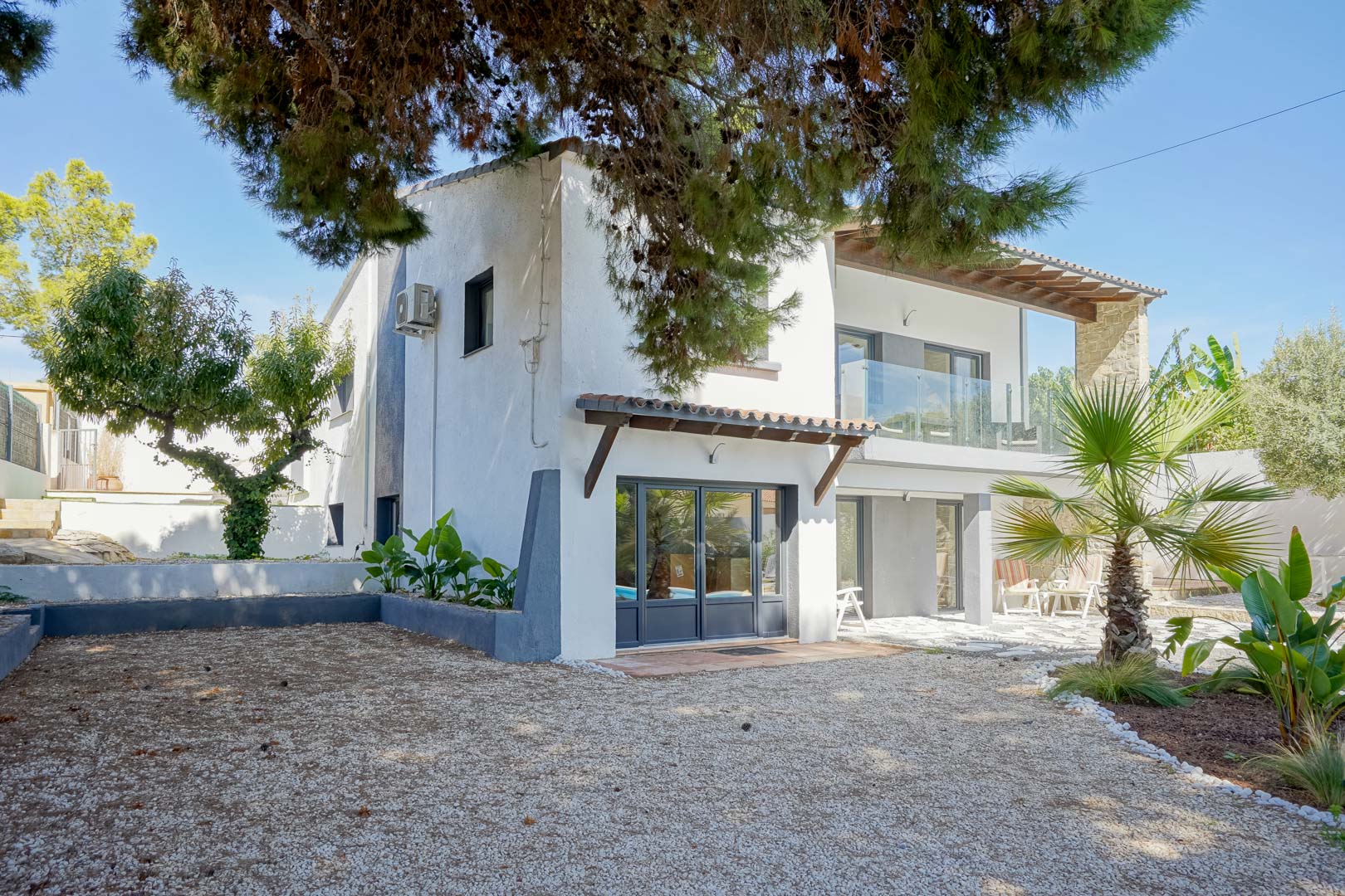 Fachada exterior de la villa con carpintería gris, terrazas con barandillas de cristal y jardín mediterráneo con grava, palmeras y pinos maduros.