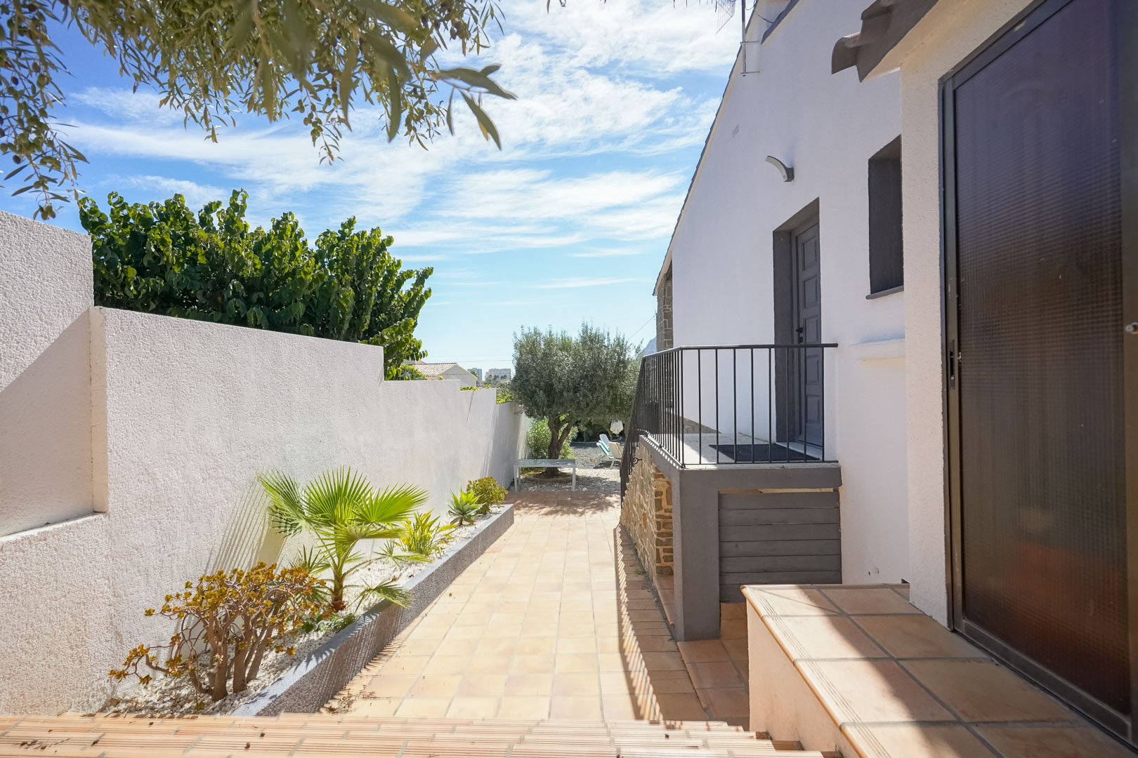 Pasillo exterior pavimentado con baldosas cerámicas, flanqueado por un muro blanco con jardineras mediterráneas y acceso lateral a la vivienda mediante escalera con barandilla metálica.