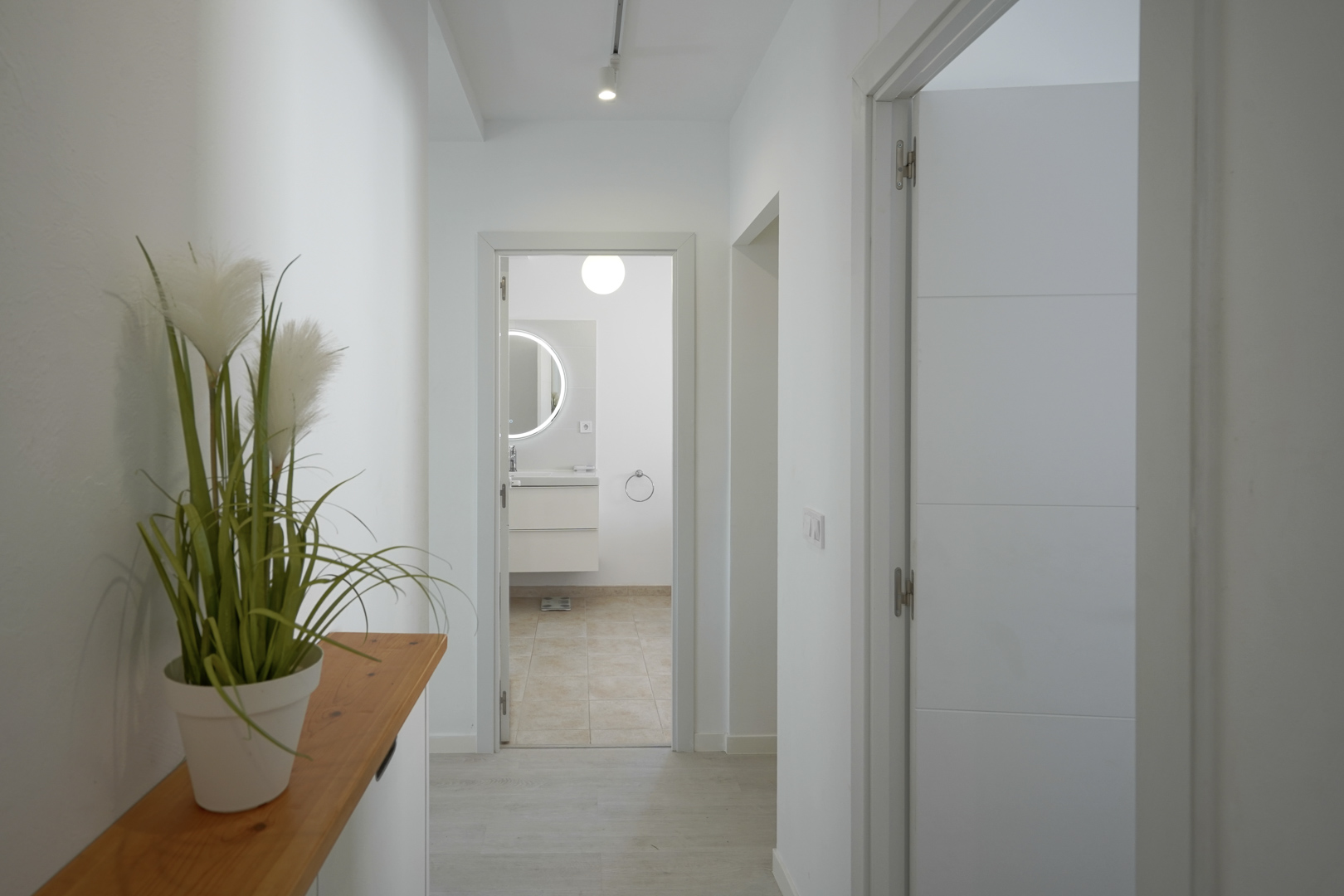 Pasillo luminoso con paredes blancas y suelos claros que conduce a un baño moderno con espejo circular retroiluminado y mueble suspendido.