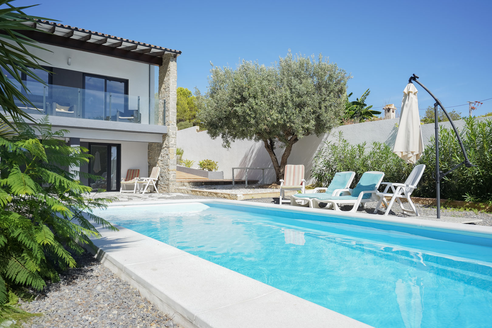 Piscina exterior rodeada de una zona de solárium con tumbonas, un olivo maduro y la fachada de la villa con balcones de cristal y detalles en piedra natural.