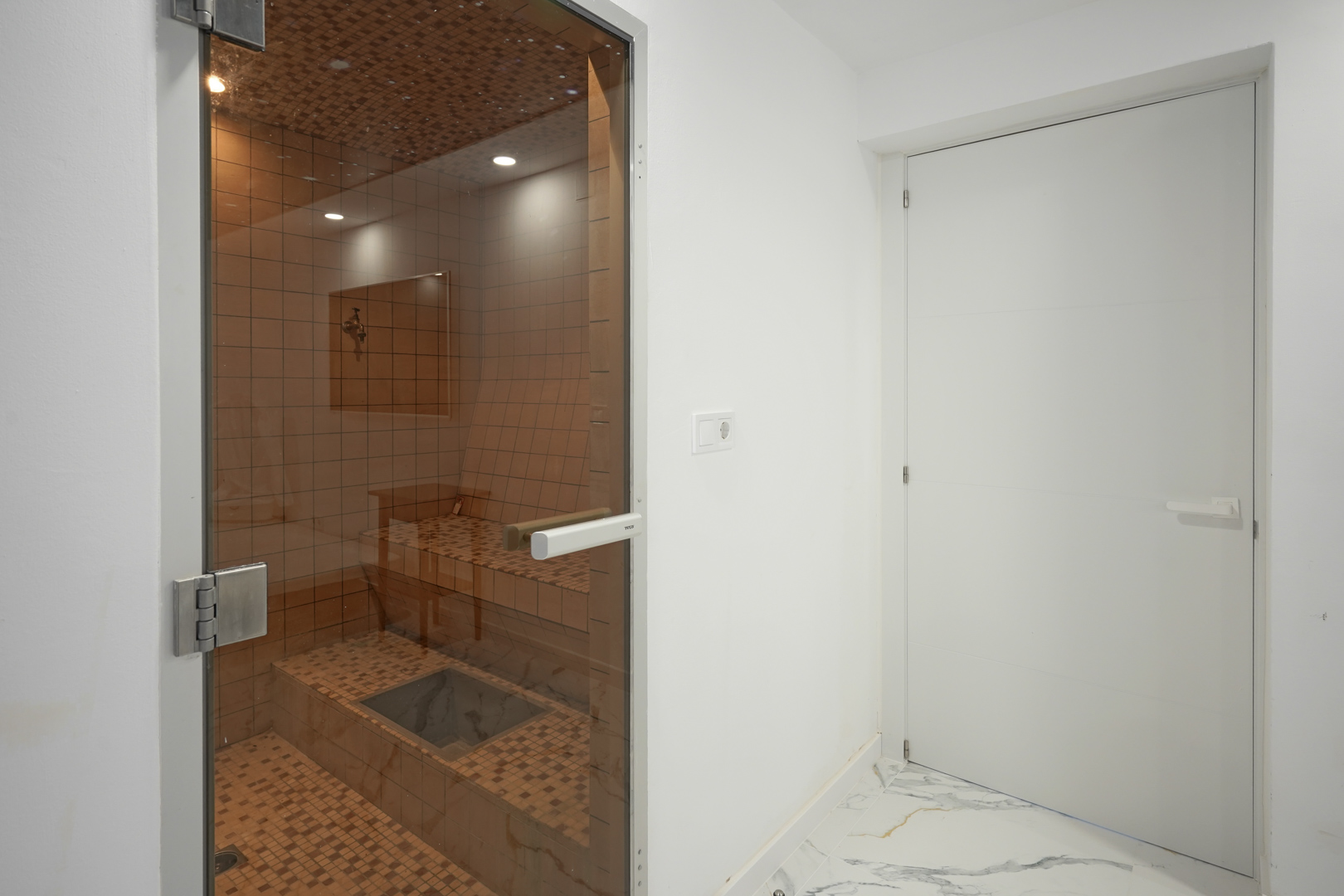 Sauna privada con revestimiento de mosaico cerámico, iluminación empotrada y puerta de cristal, integrada en una estancia con suelos de mármol.