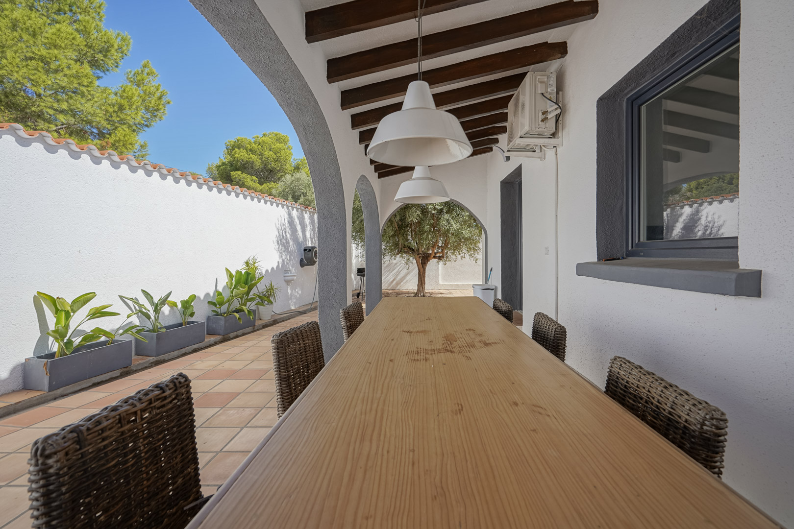 Terraza cubierta con vigas de madera, mesa de comedor alargada y sillas de mimbre. El espacio cuenta con arcos mediterráneos y vistas al jardín con olivos.