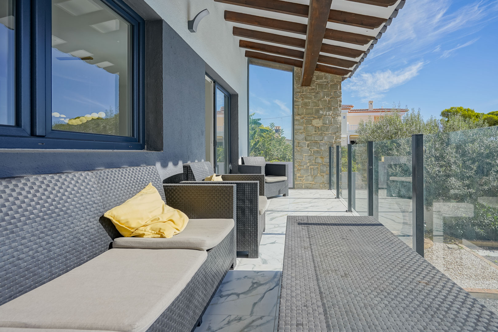 Terraza cubierta con vigas de madera, mobiliario de ratán sintético y suelo de mármol. Incluye barandilla de cristal y vistas despejadas al entorno mediterráneo.