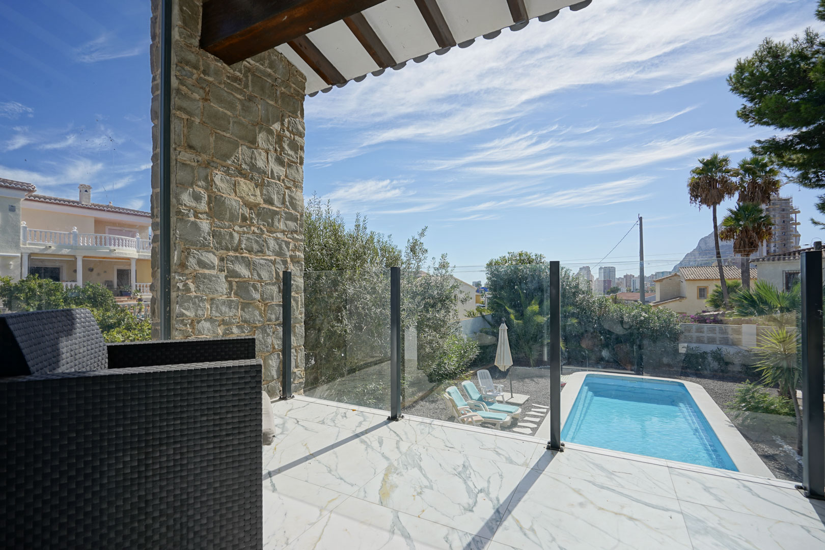 Terraza superior con suelo de mármol, barandilla de cristal y vistas a la piscina privada, el jardín mediterráneo y el Peñón de Ifach al fondo.