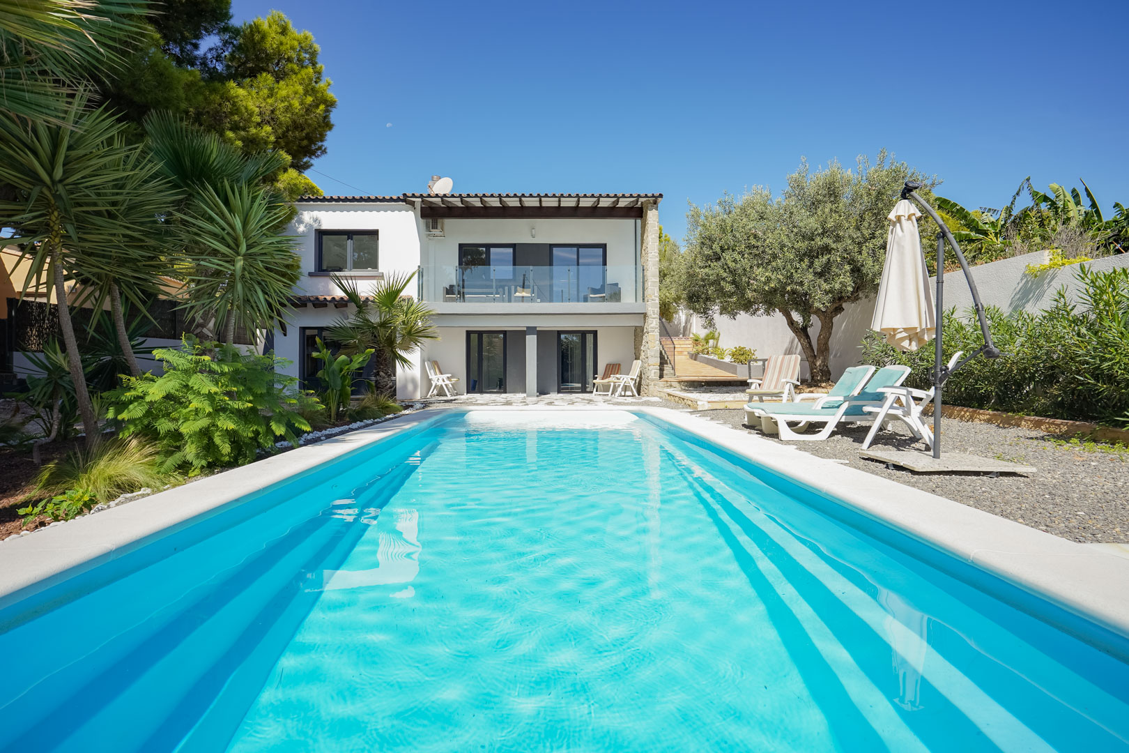 Vista de la piscina rectangular y jardín mediterráneo con palmeras, destacando la fachada blanca de la villa y su terraza superior con barandilla de cristal.