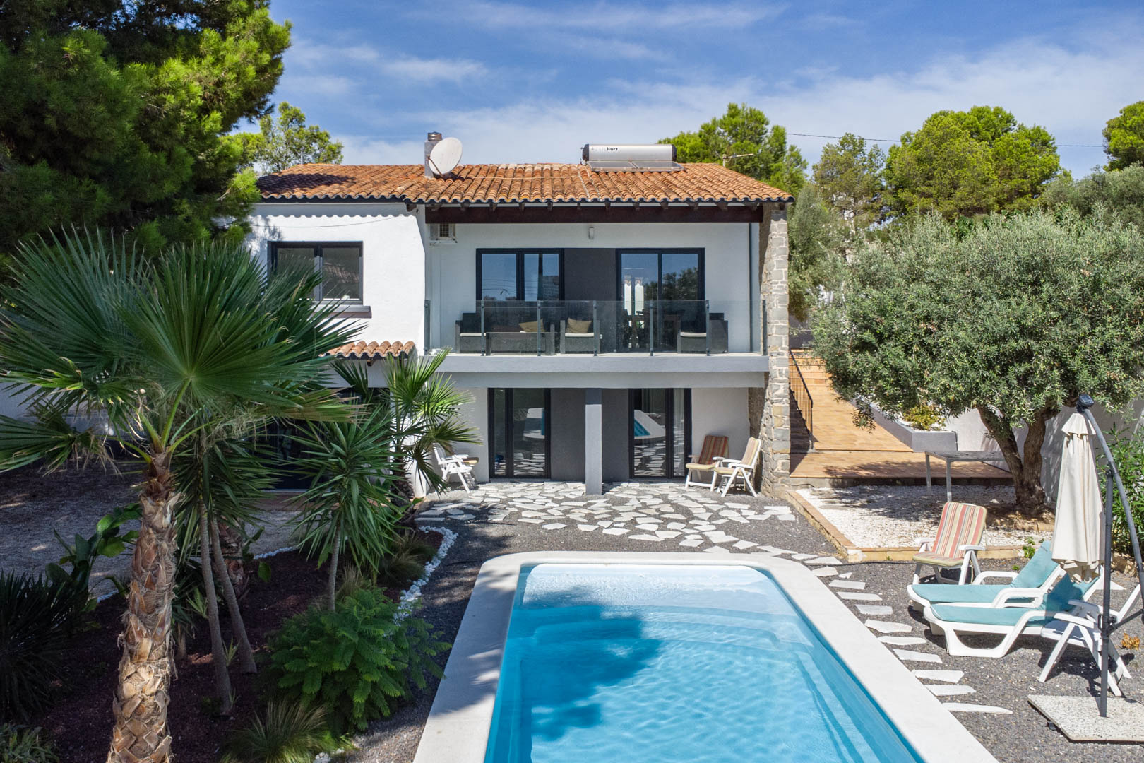 Vista exterior de la villa con piscina rectangular, jardín mediterráneo y fachada blanca. Incluye terrazas con barandillas de cristal y calentador solar en el tejado.