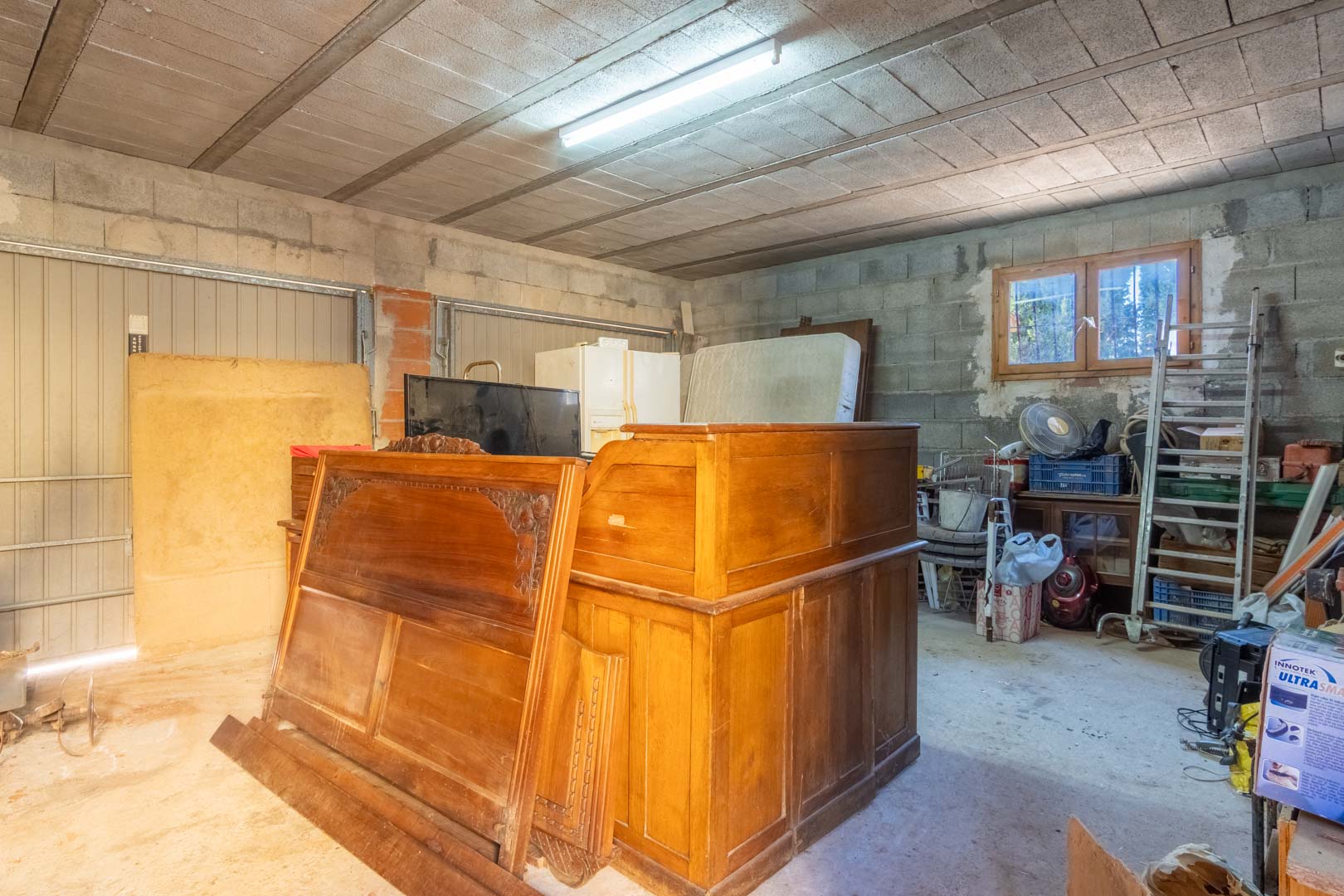 Amplio trastero o garaje con paredes de bloque de hormigón, techos de viguetas y bovedillas, iluminación fluorescente y luz natural a través de una ventana de madera.