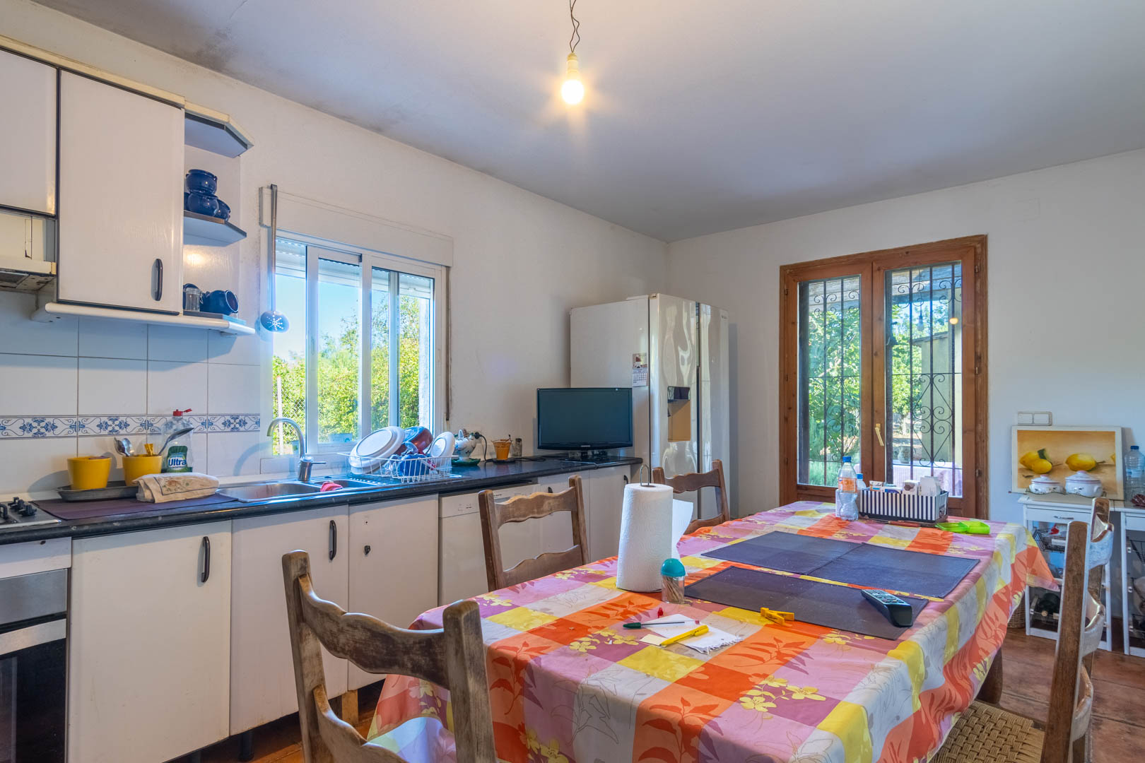 Cocina funcional con armarios blancos, encimera oscura y zona de comedor. Incluye acceso directo al exterior y vistas al jardín a través de ventanales con carpintería de madera.