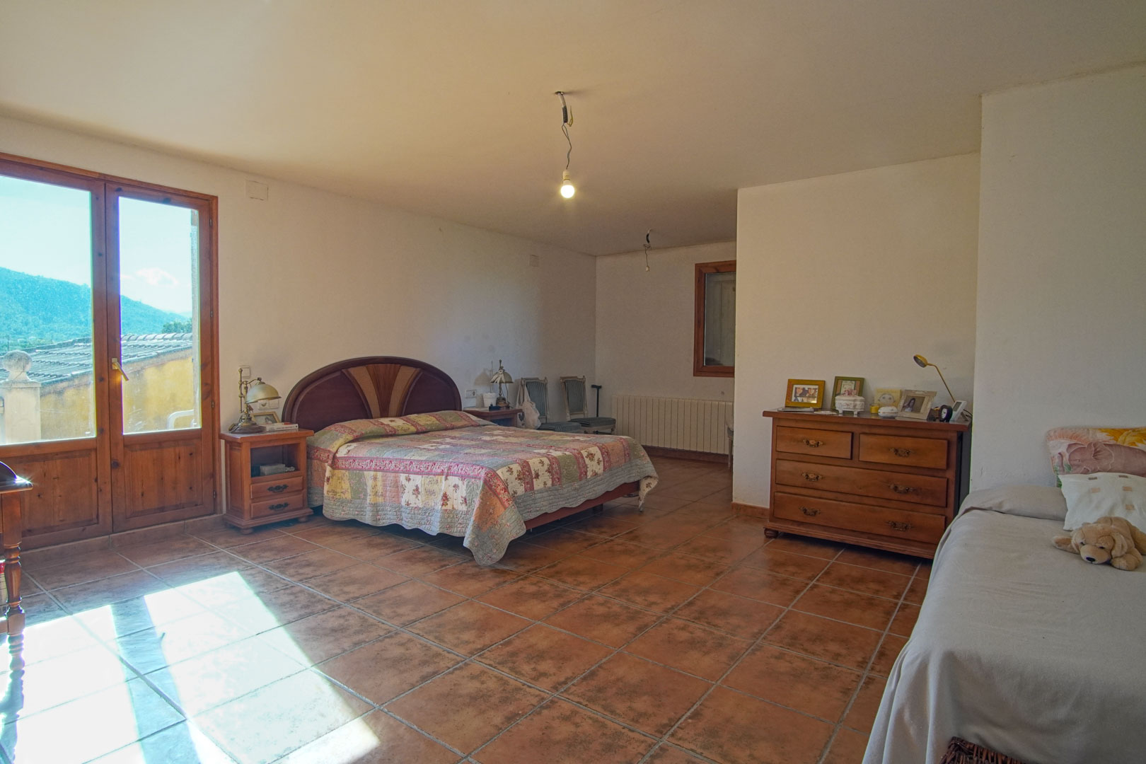 Dormitorio amplio con suelo de gres rústico, mobiliario de madera clásica y acceso directo a través de una puerta acristalada con vistas a la montaña.