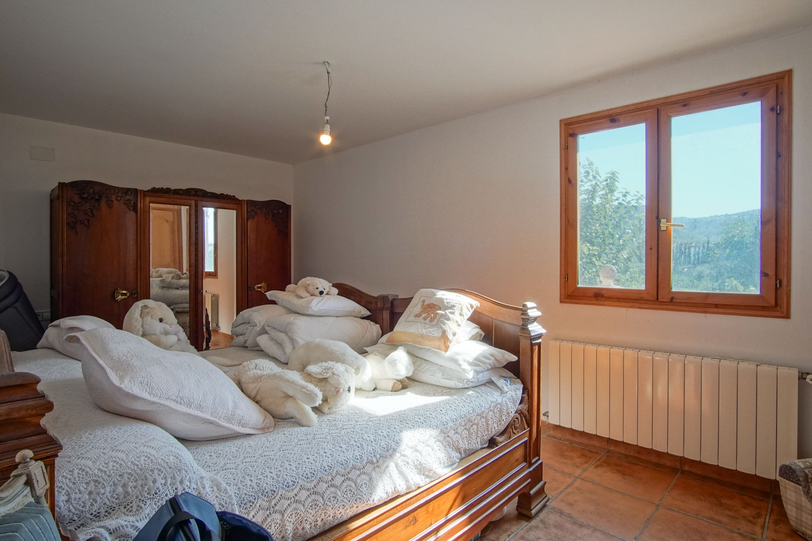 Dormitorio con mobiliario de madera tallada, armario con espejos, suelos de terracota y ventana con vistas a la montaña y calefacción por radiador.