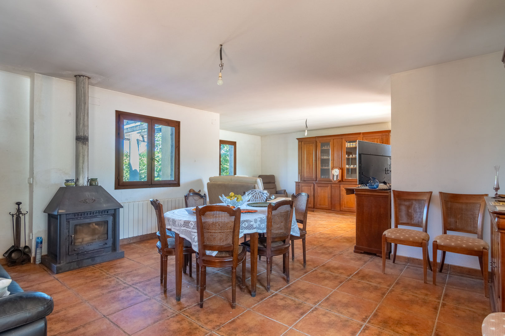 Salón-comedor con suelos de gres rústico, presidido por una chimenea de leña metálica y mobiliario de madera clásica con vitrina de gran capacidad.