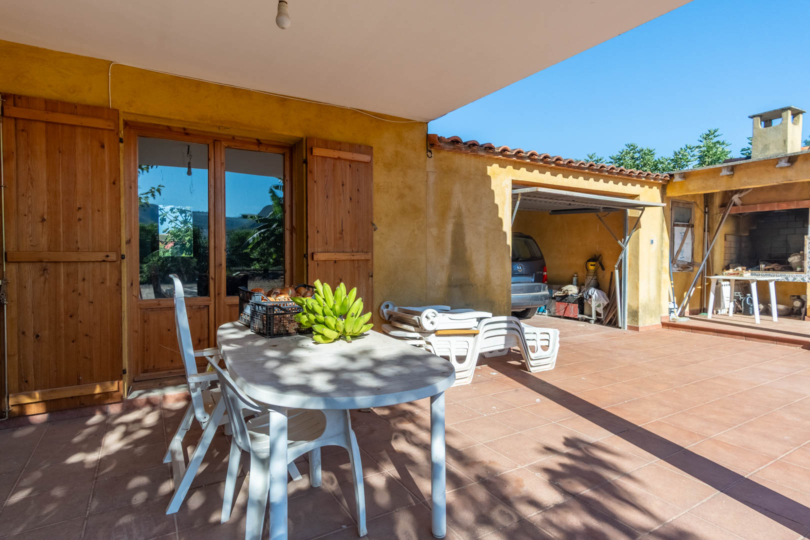 Terraza exterior con suelo de gres cerámico, zona de comedor bajo porche y acceso a garaje cubierto y paellero tradicional con barbacoa.