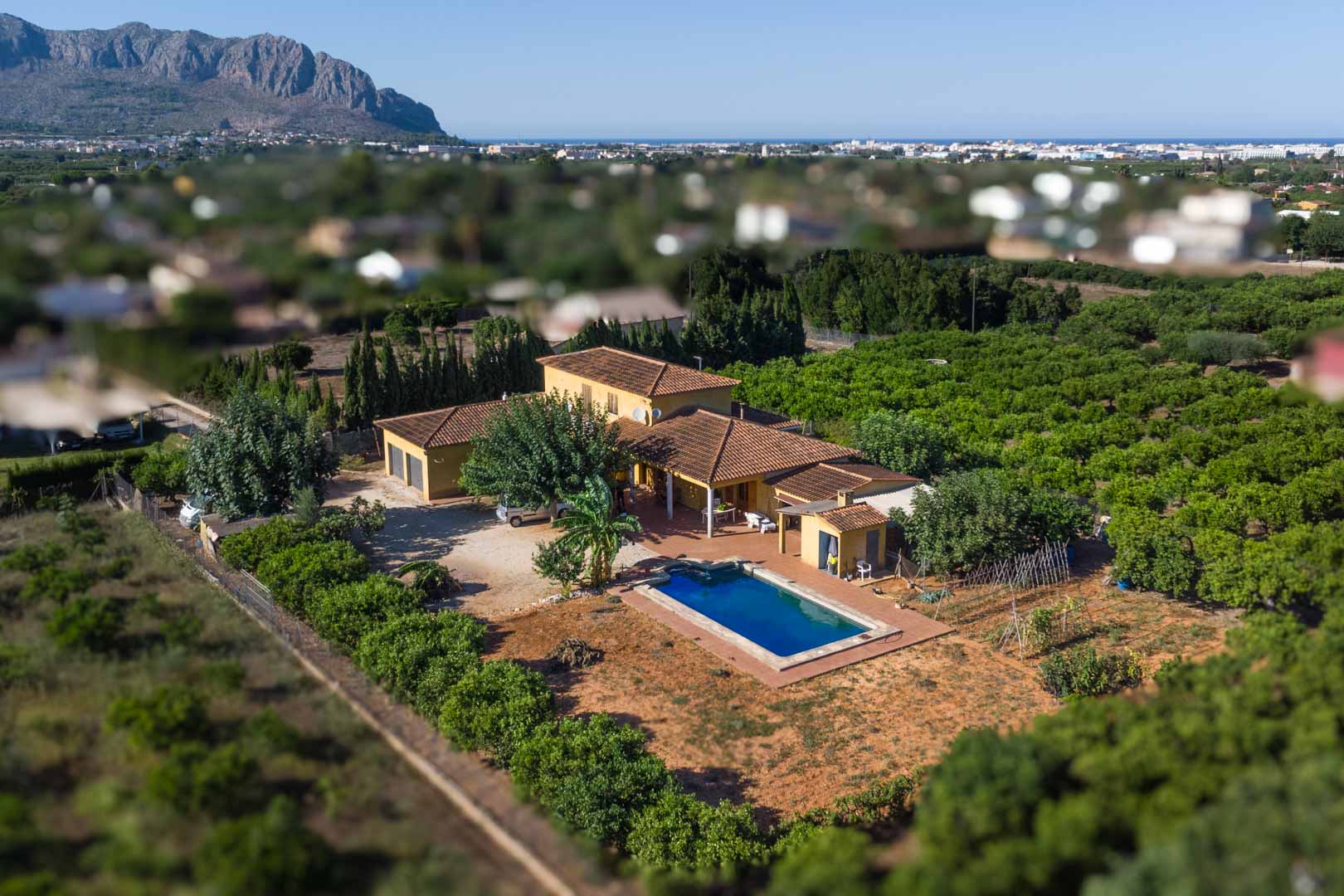 Vista aérea de la villa con piscina rectangular, rodeada de huertos y vegetación mediterránea, con la montaña Segaria y la costa en el horizonte.