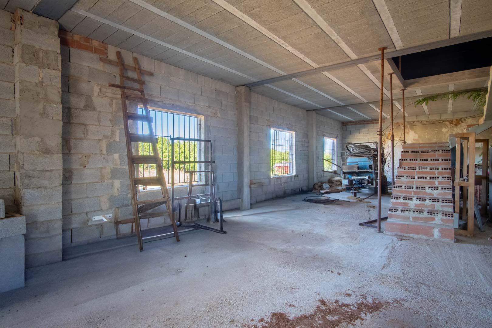 Espacioso interior diáfano en fase de reforma con muros de bloque, forjado de viguetas y ventanales con rejas que aportan luz natural y vistas al entorno rural.