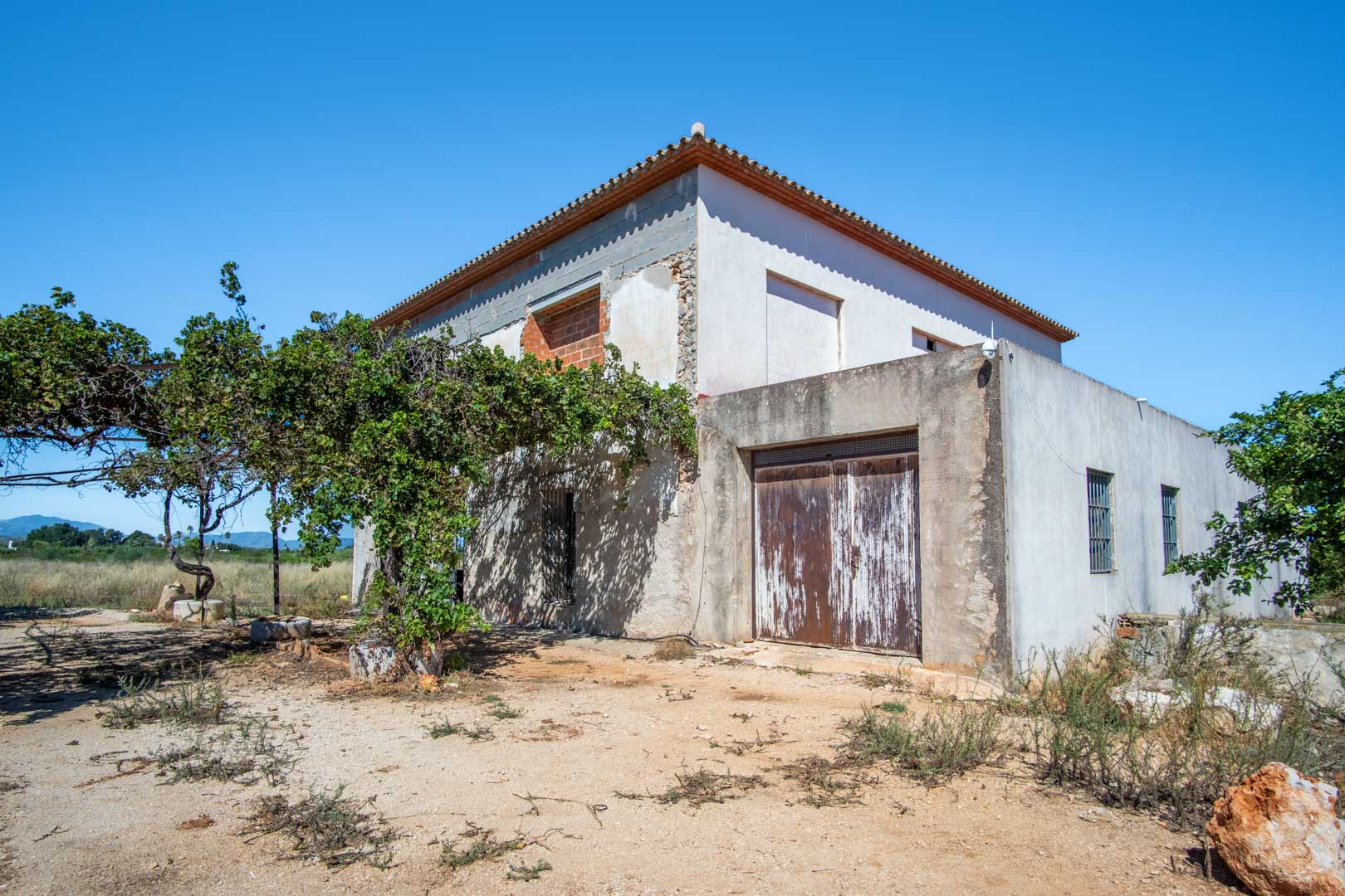 Fachada exterior de la casa de campo en proceso de reforma, mostrando la estructura consolidada, el terreno llano y vistas despejadas a las montañas de Dénia.