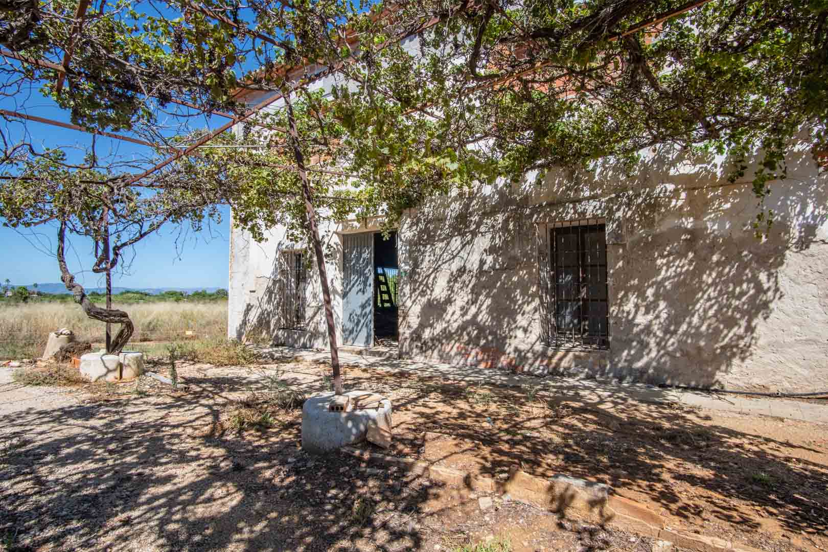 Fachada rústica con estructura de parra natural que proporciona sombra a la entrada principal, destacando los muros blancos y las ventanas con rejería tradicional.