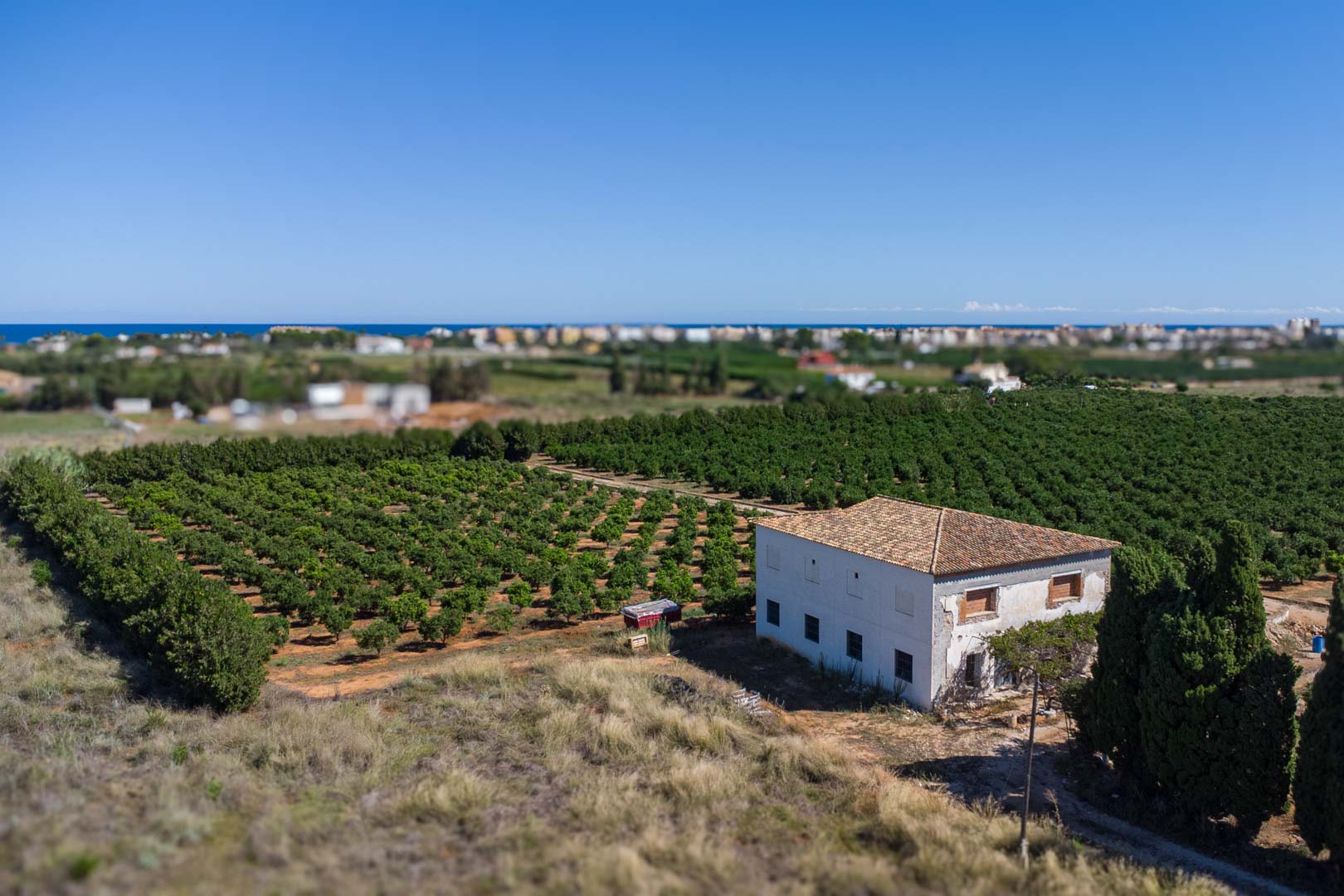 Vista aérea de la casa de campo rodeada de cultivos, con fachada blanca, tejado de teja y vistas panorámicas al mar Mediterráneo en el horizonte.