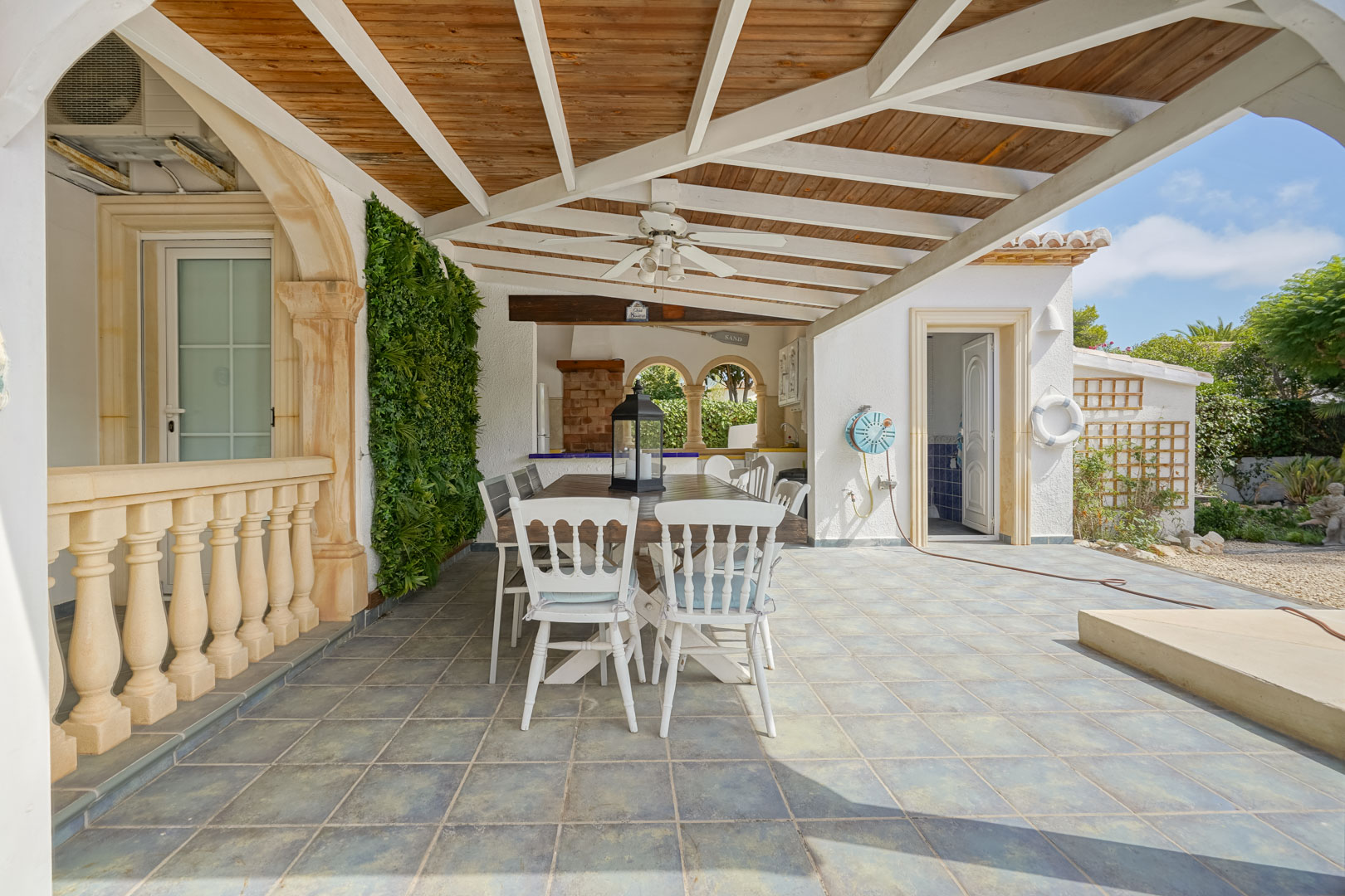 Comedor exterior cubierto con mesa de madera y sillas blancas, junto a una cocina exterior con barbacoa.
